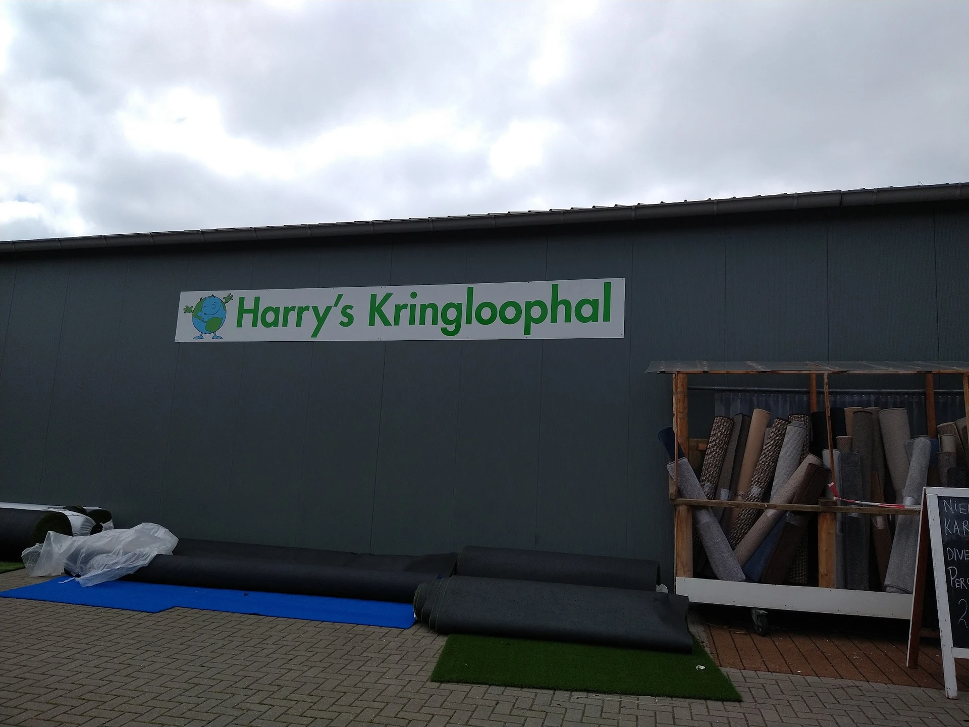 Gevel van kringloopwinkel met groen logo 'Harry's Kringloophal' op donkere muur, daaronder blauwe en groene matten en materia