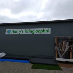 Gevel van kringloopwinkel met groen logo 'Harry's Kringloophal' op donkere muur, daaronder blauwe en groene matten en materia