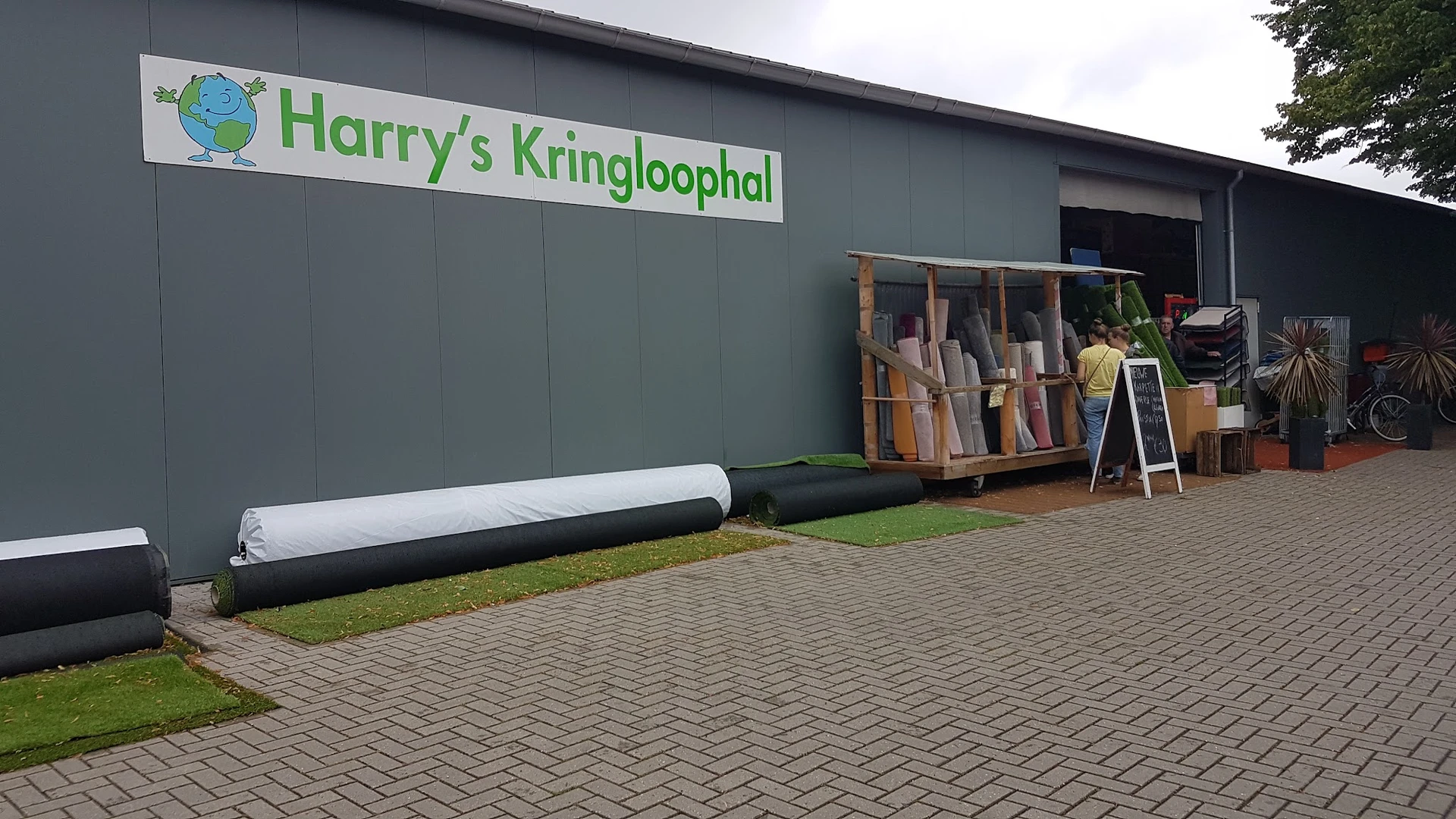 Grijze gevel van Harry's Kringloophal met groen logo, voorzijde met houten materialen en bouwmateriaal op stapel voor de inga