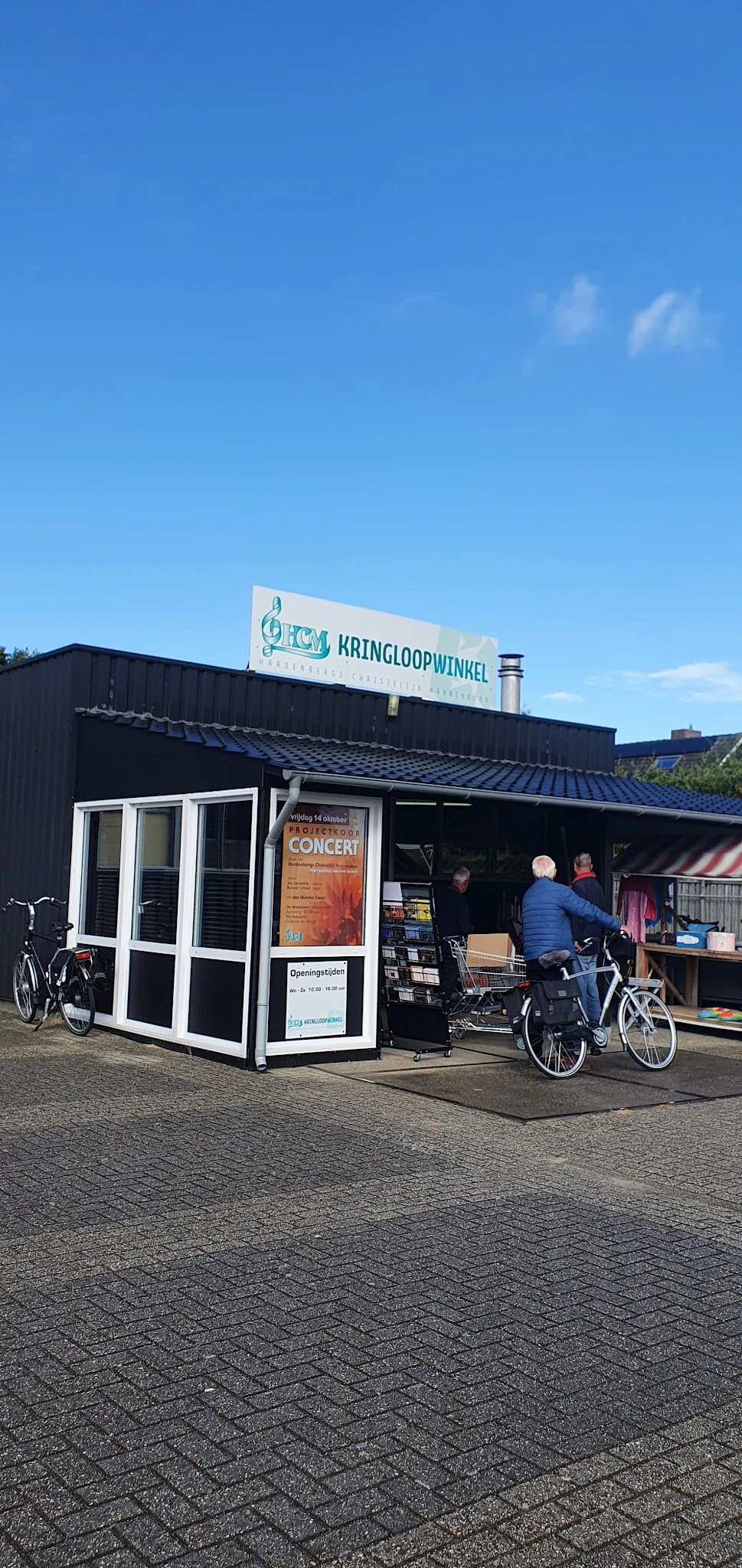 Extern zwart gebouw met witte deur en raam, winkelnaam op bord, fietsen ervoor, oranje concert-poster zichtbaar