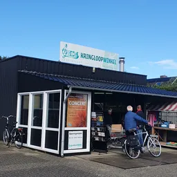 Extern zwart gebouw met witte deur en raam, winkelnaam op bord, fietsen ervoor, oranje concert-poster zichtbaar