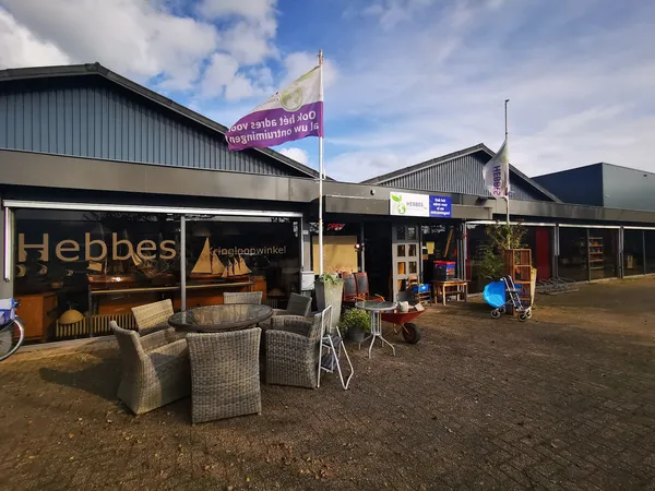Voorkant van kringloopwinkel Hebbes met naam op raam, terras met stoelen en personeel in deuropening