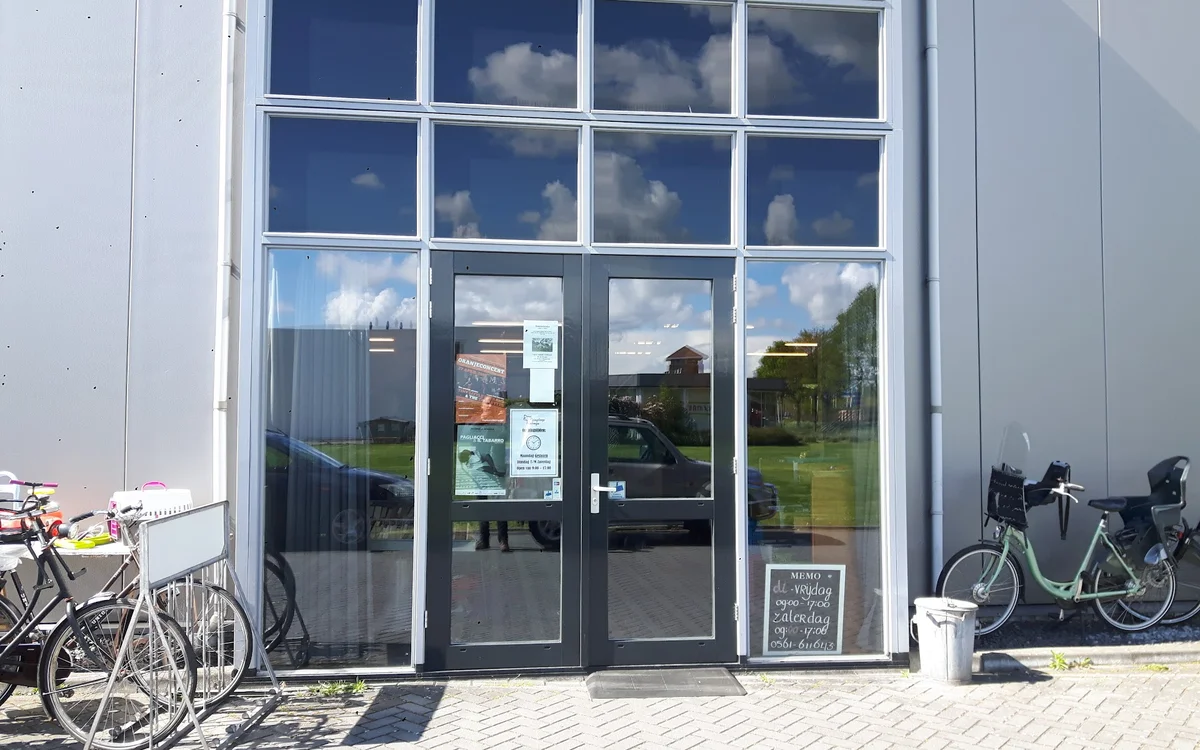 Glazen voorgevel met dubbele deuren van een kringloopwinkel, met fietsen geparkeerd aan weerskanten