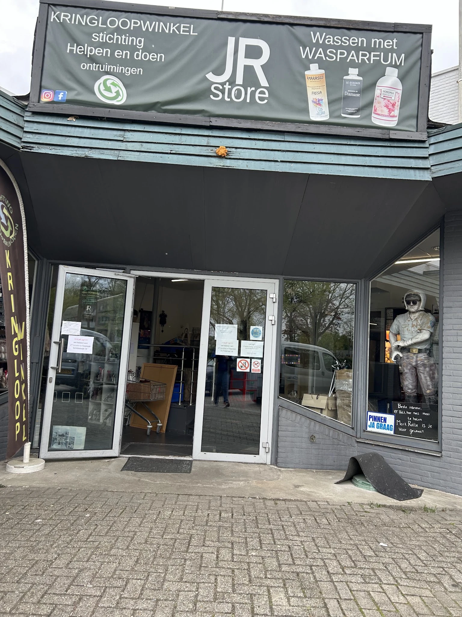 Voorgevel van kringloopwinkel met glazen deuren en etalages, onderkant van uithangbord zichtbaar