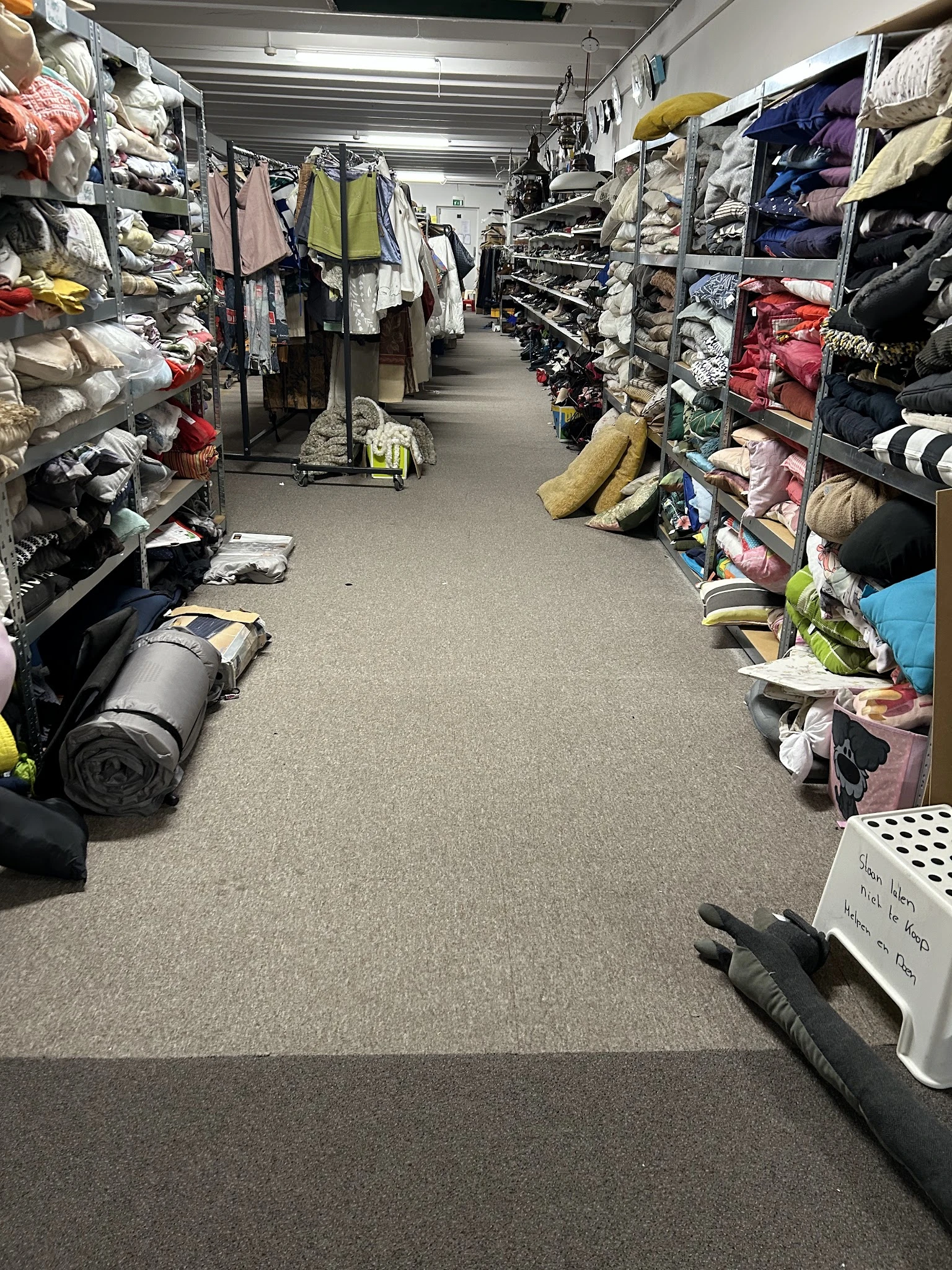 Winkelgang met stellingen vol kleding, schoenen en huishoudspullen aan beide zijden