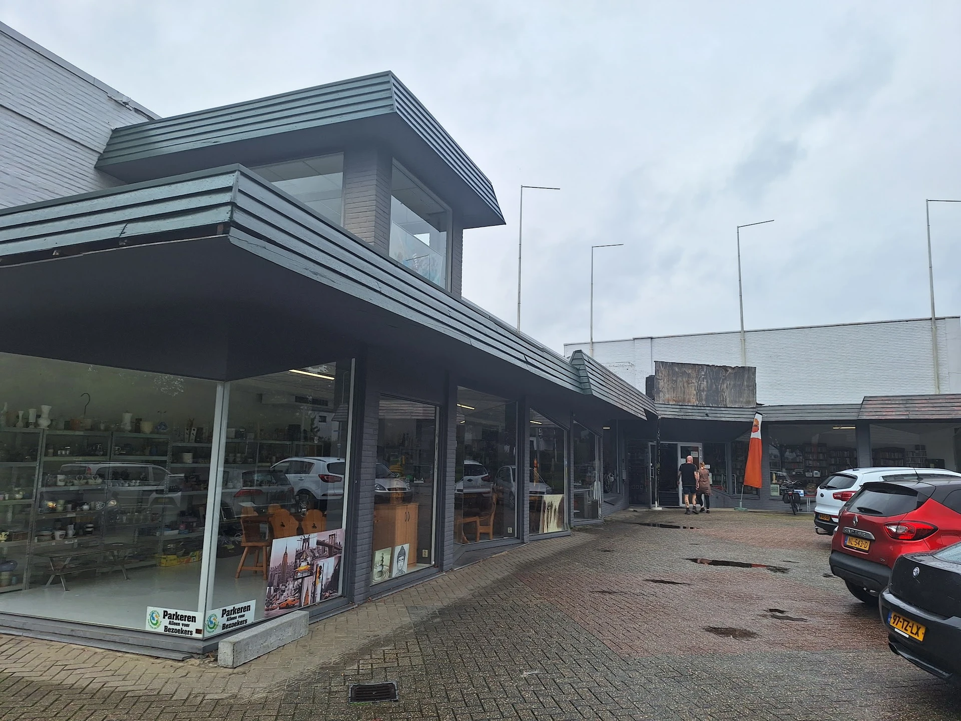 Voorgevel van kringloopwinkel met etalage, grijze bakstenen gevel en moderne lijsten, parkeerplaats met auto's.