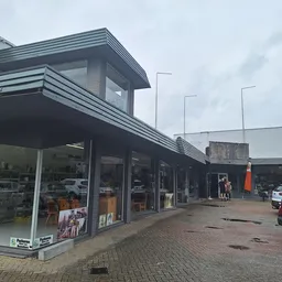 Voorgevel van kringloopwinkel met etalage, grijze bakstenen gevel en moderne lijsten, parkeerplaats met auto's.