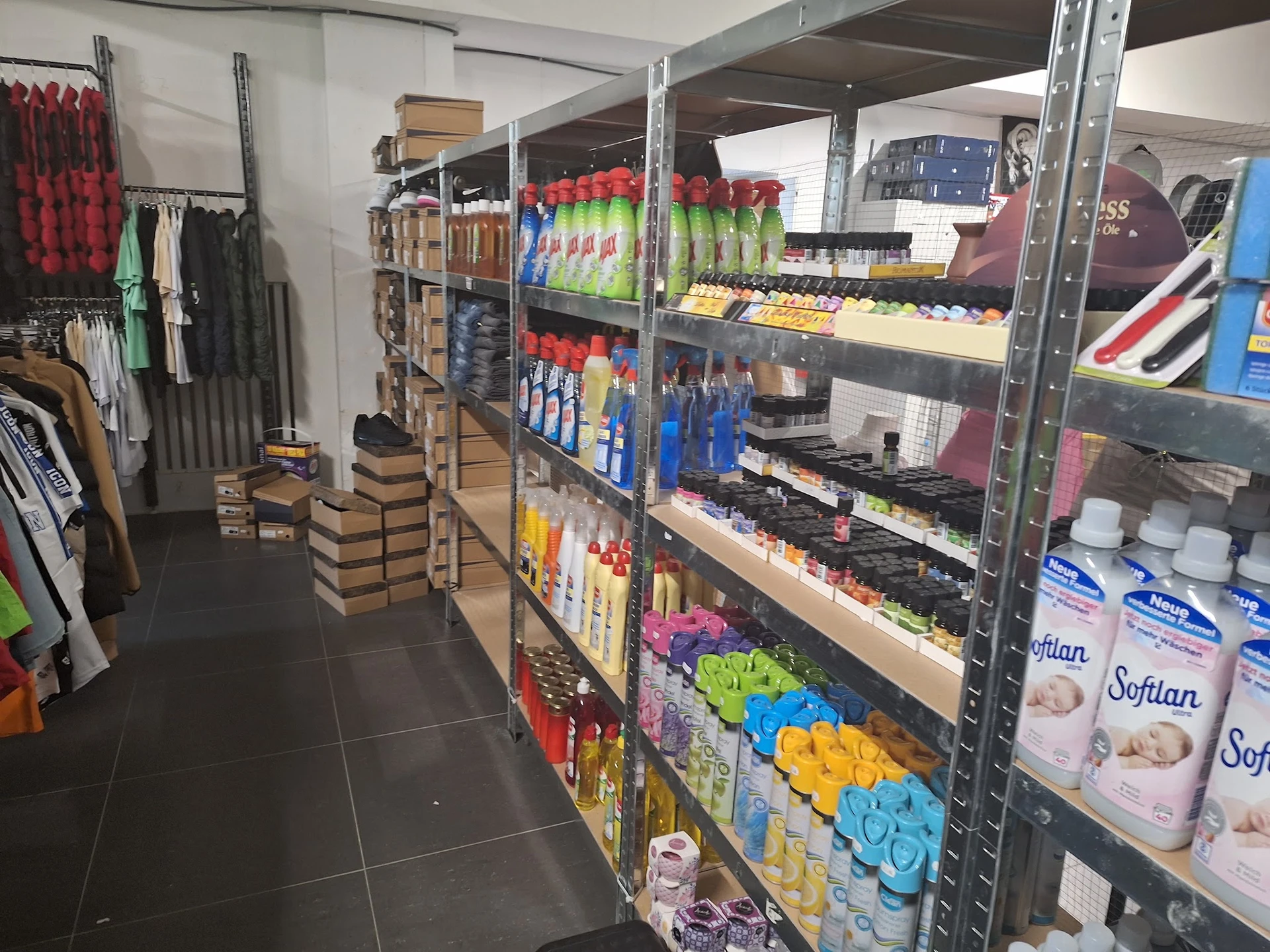 Winkelgang met metalen stellingen vol huishoudproducten, schoonmaakmiddelen en textiel, links kleding aan rekken