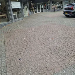 Straatscène met geplaveide voetpad, winkelpuien links en geparkeerde auto rechts