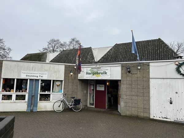 Voorgevel van kringloopwinkel Hergebruik met witte puien, rode deur en twee dakkapellen, fiets geparkeerd ervoor