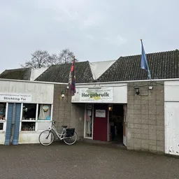 Voorgevel van kringloopwinkel Hergebruik met witte puien, rode deur en twee dakkapellen, fiets geparkeerd ervoor