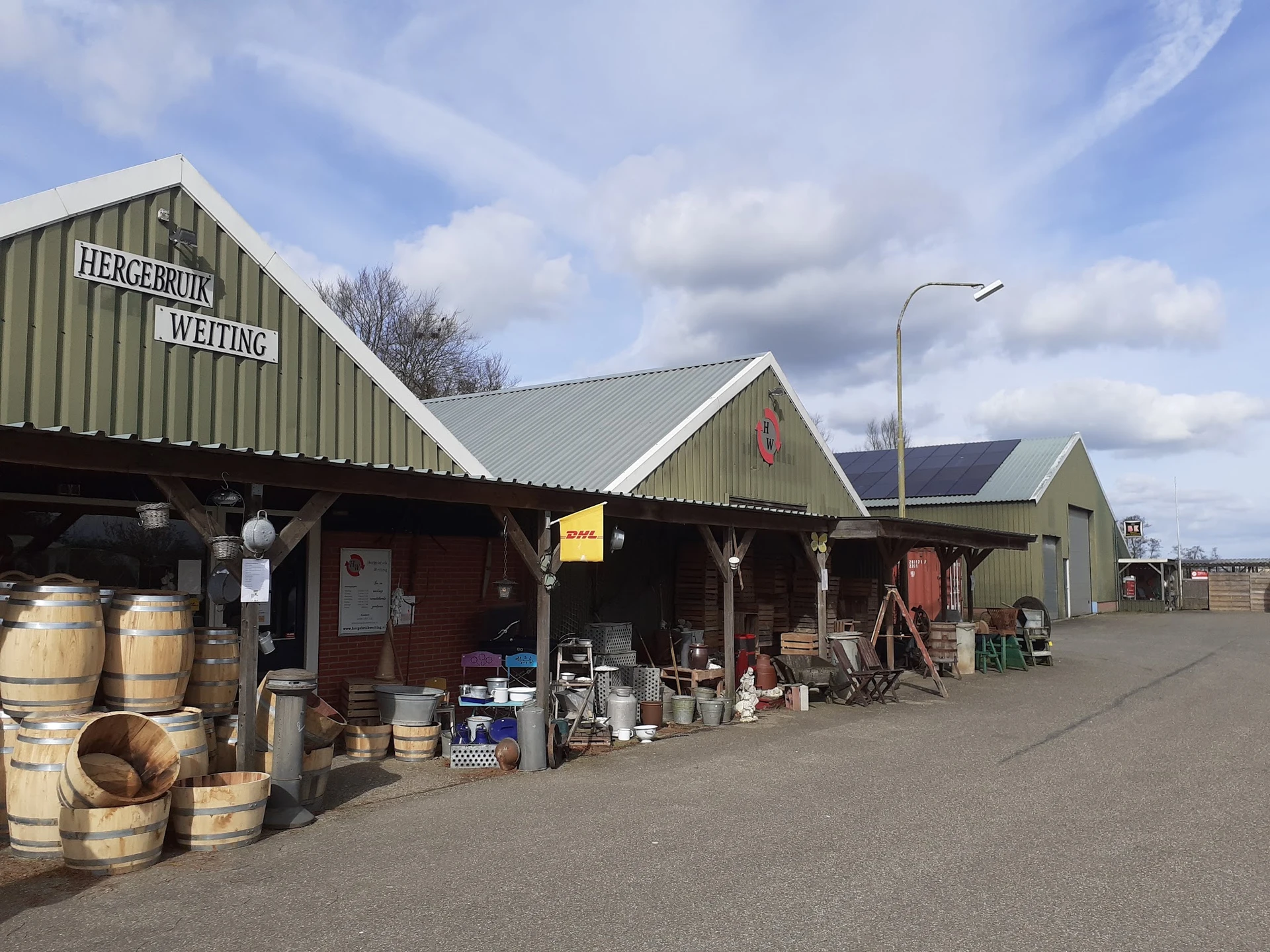 Voorgevel van kringloopwinkel met groene daken, houten vaten en diverse spullen buiten, aan de straat