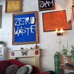Interieur van kringloopwinkel met aan muur opgehangen kunstwerken, waaronder blauw schilderij met 'ZERO WASTE' tekst, decorat