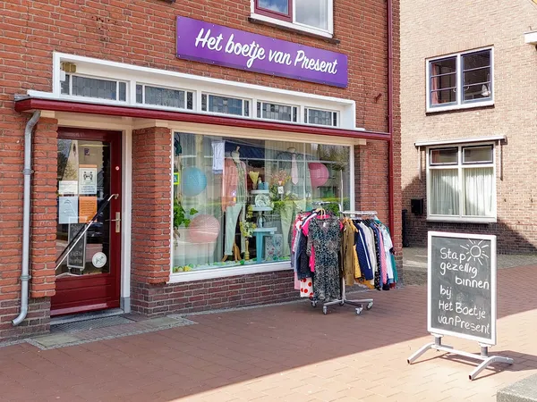Voorkant van kringloopwinkel met paarse naambordje, etalage met kleding op rekken en winkelzijkant met stoepbord