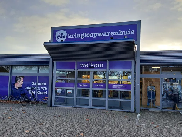 Gevel van kringloopwinkel met blauwe pui, winkelnaam op groot bord en welkomsignage op ramen