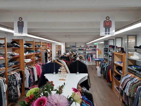 Winkelinterieur met houten stellingen vol tweedehands kleding, bloemen op een toonbank en geslachtsaanduidingen boven.