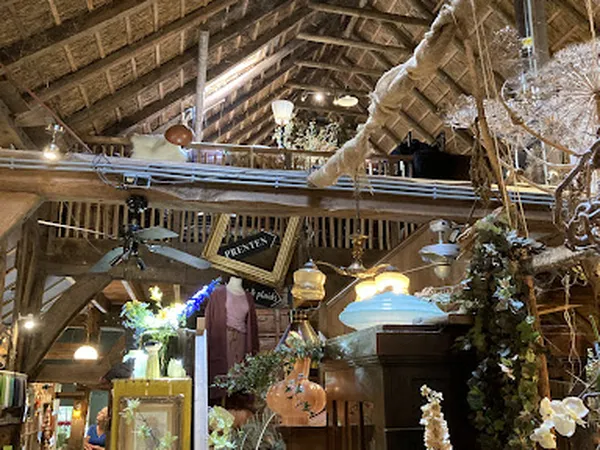 Binnenaanzicht van een kringloopwinkel met houten balken, hanglampen en diverse spullen zoals bloemen, kunstwerken en decorat