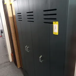 Zwarte kasten of servers met ventilatiesleuven en een geel etiket tegen een muur.