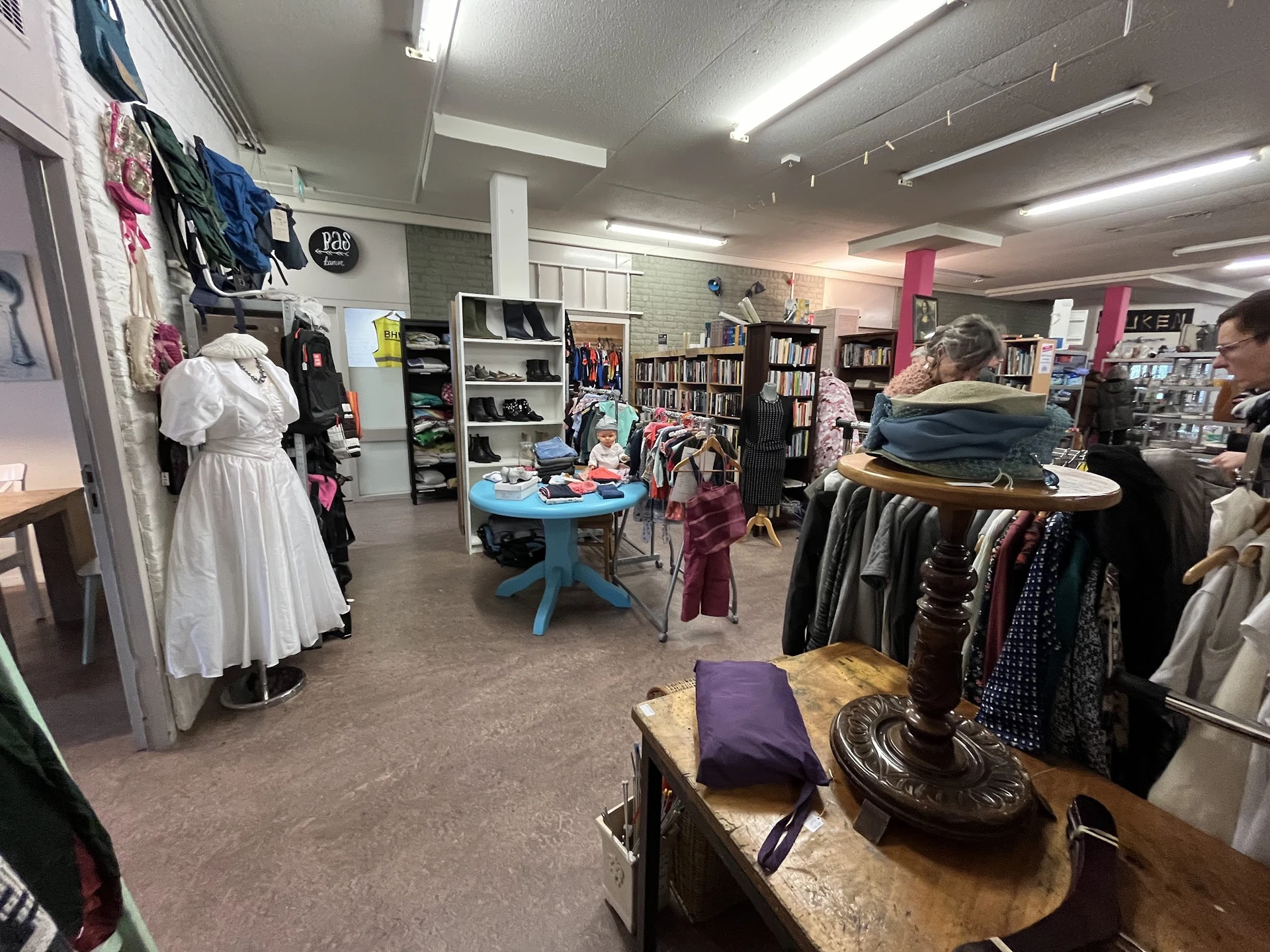 Winkelvloer met kledingrekken, boekenkasten, blauwe tafel en bezoekers in een kringloopwinkel