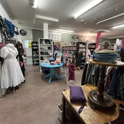 Winkelvloer met kledingrekken, boekenkasten, blauwe tafel en bezoekers in een kringloopwinkel