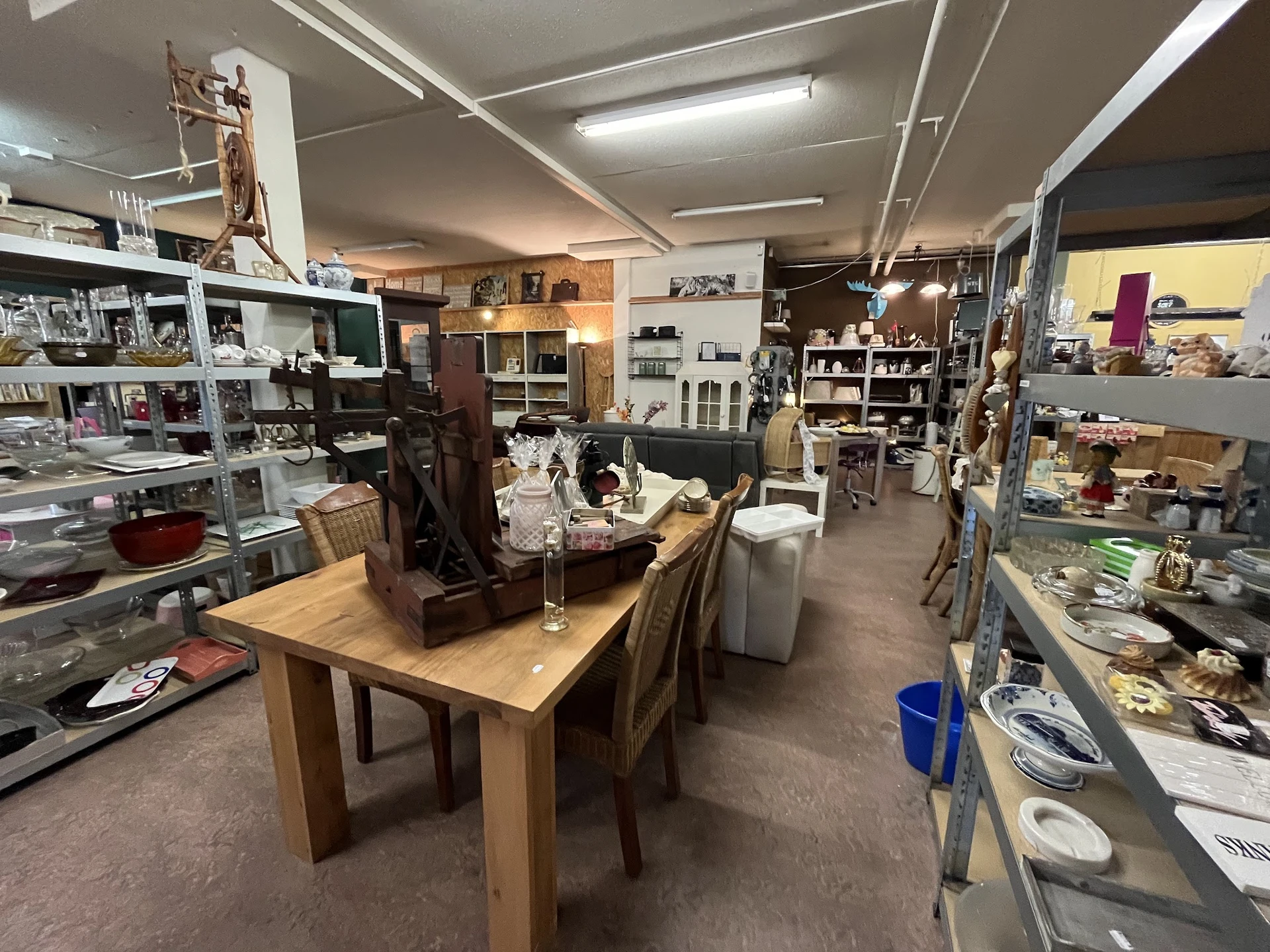 Ruim kringloopwinkelinterieur met houten tafel, stellingen met artikelen, verlichting en diverse inrichtingsspullen