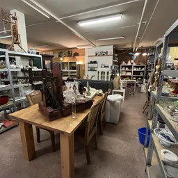 Ruim kringloopwinkelinterieur met houten tafel, stellingen met artikelen, verlichting en diverse inrichtingsspullen