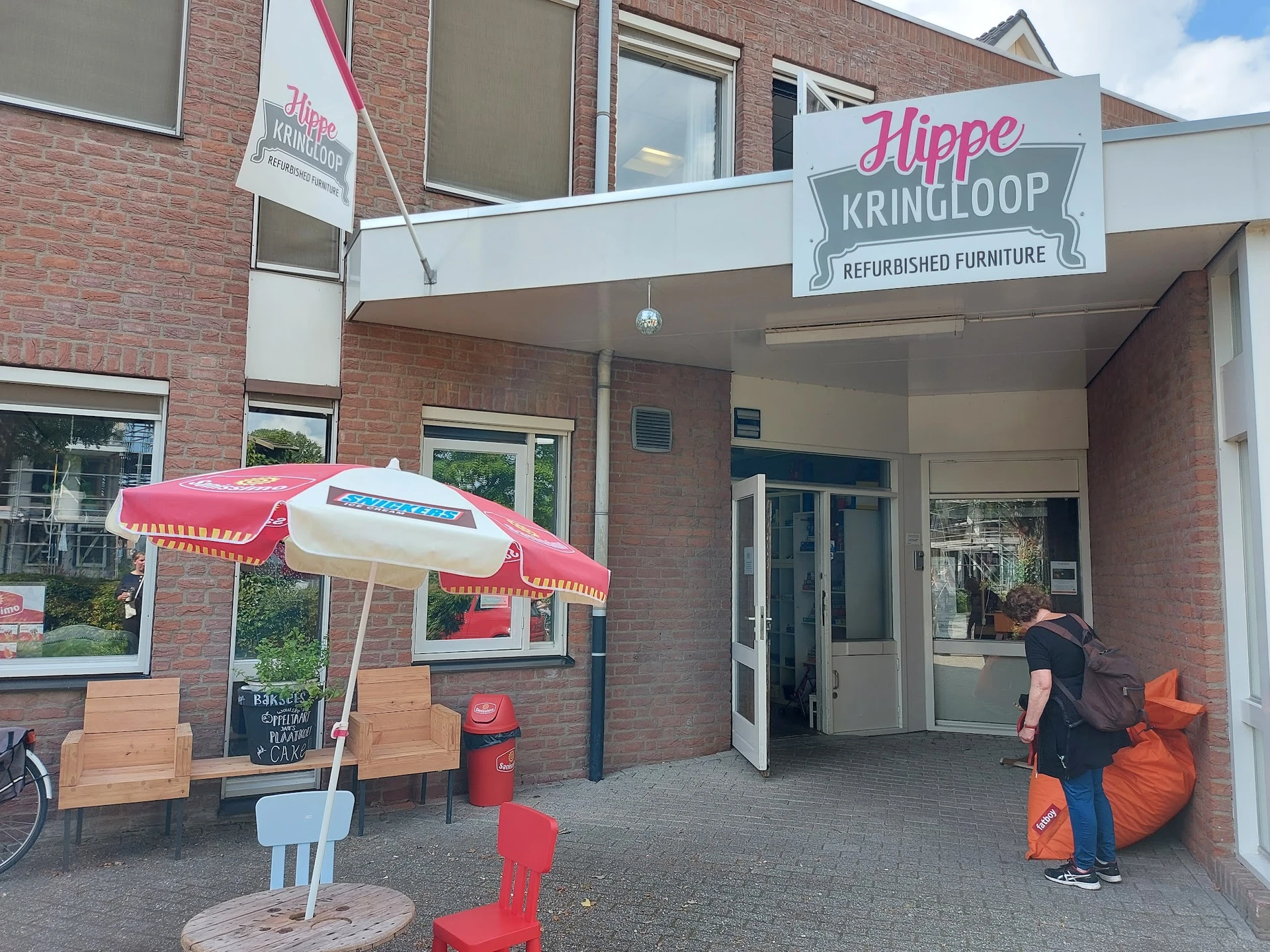 Ingang van Hippe Kringloop met terras, rode parasol, stoelen en oranje poefjes