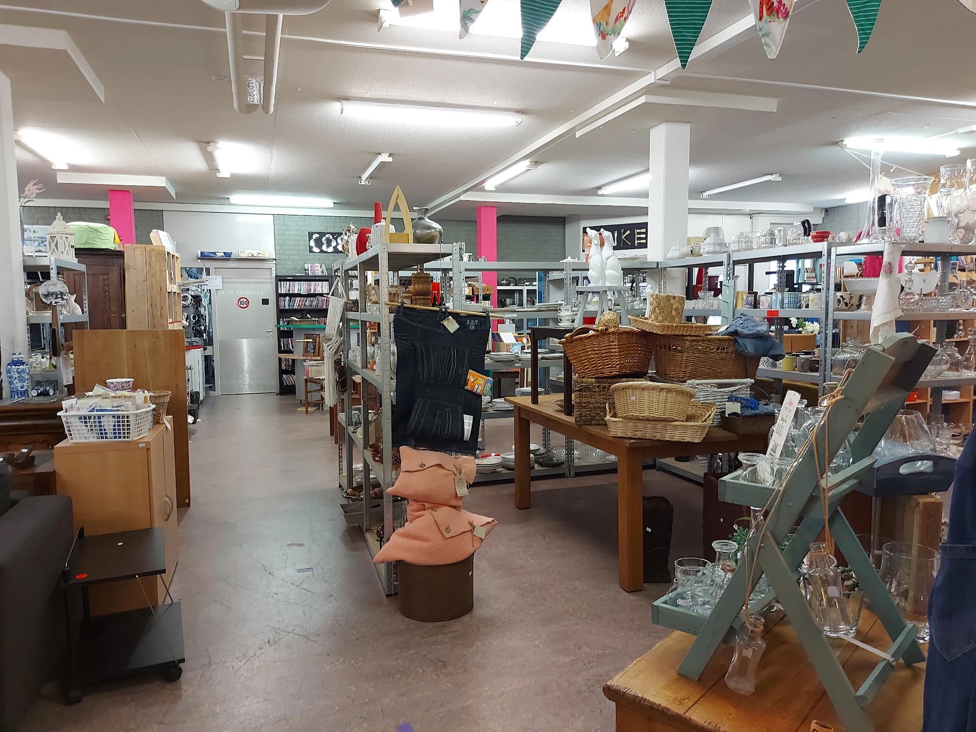 Ruime winkelvloer van kringloopwinkel met meubels, dozen en spullen verspreid uitgestald onder fluorescente verlichting