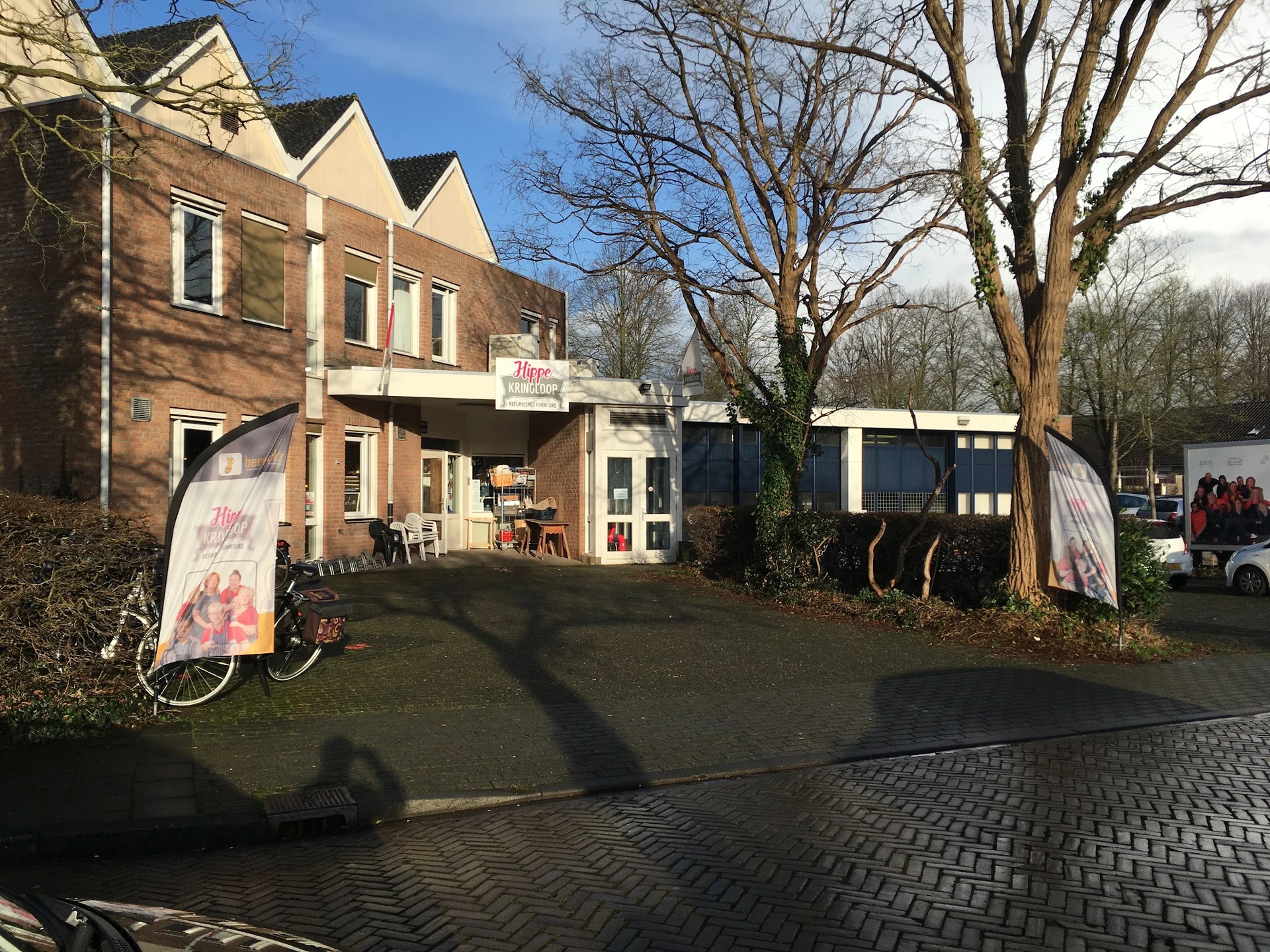 Ingang van kringloopwinkel met bruine bakstenen gevel, witte deurkozijnen en informatieborden aan de voorkant