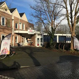 Ingang van kringloopwinkel met bruine bakstenen gevel, witte deurkozijnen en informatieborden aan de voorkant