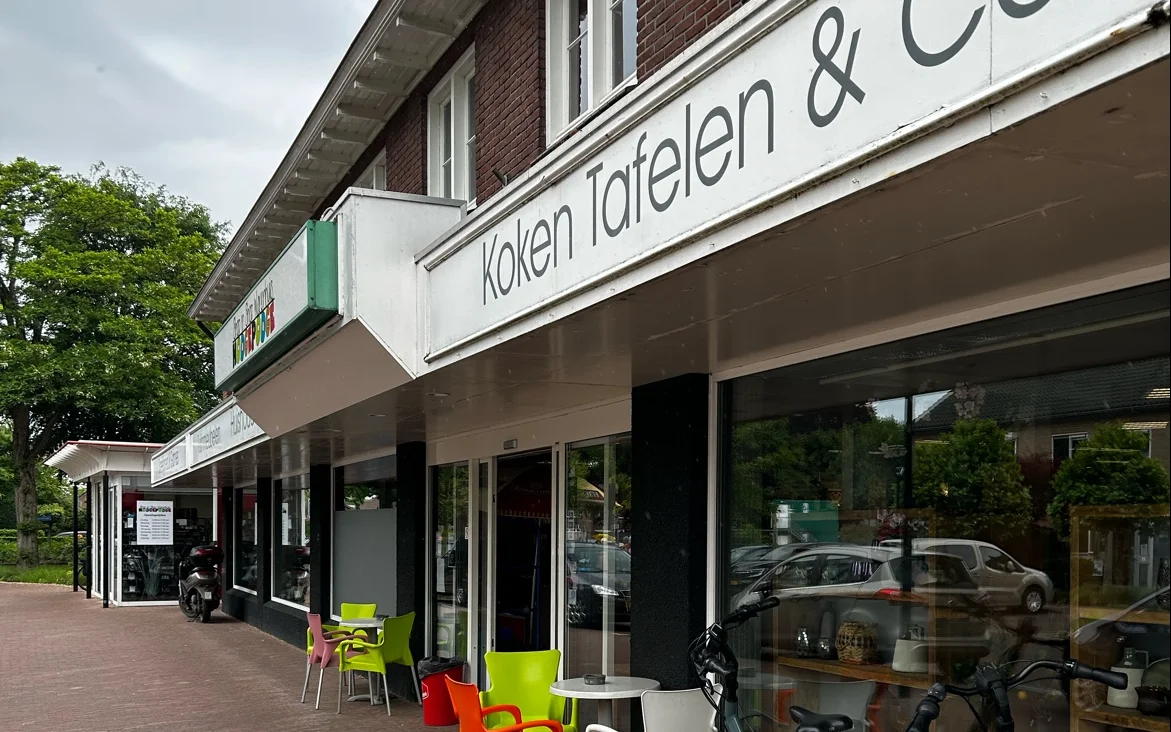Winkelgevel met witte markiezen en groene accenten, uitstalramen met fietsen en artikelen, zitjes op het terras.