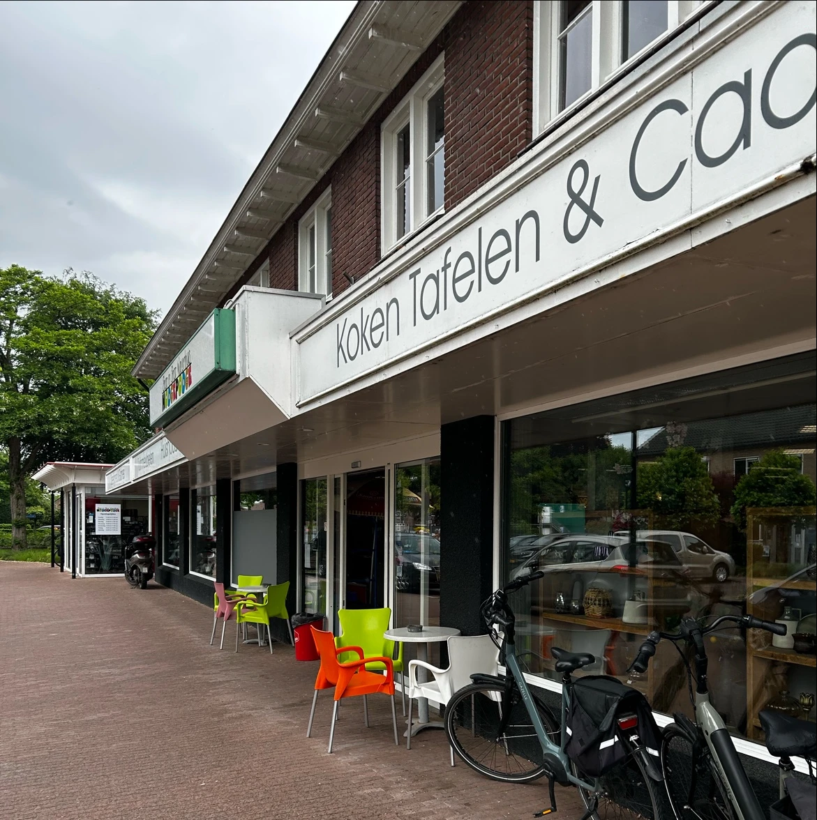 Winkelgevel met witte markiezen en groene accenten, uitstalramen met fietsen en artikelen, zitjes op het terras.