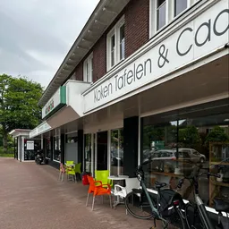 Winkelgevel met witte markiezen en groene accenten, uitstalramen met fietsen en artikelen, zitjes op het terras.
