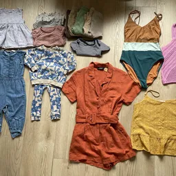 Assortiment tweedehands kleding uitgestald op houten vloer: jurken, jumpsuits, badpakken en tops in verschillende kleuren en 