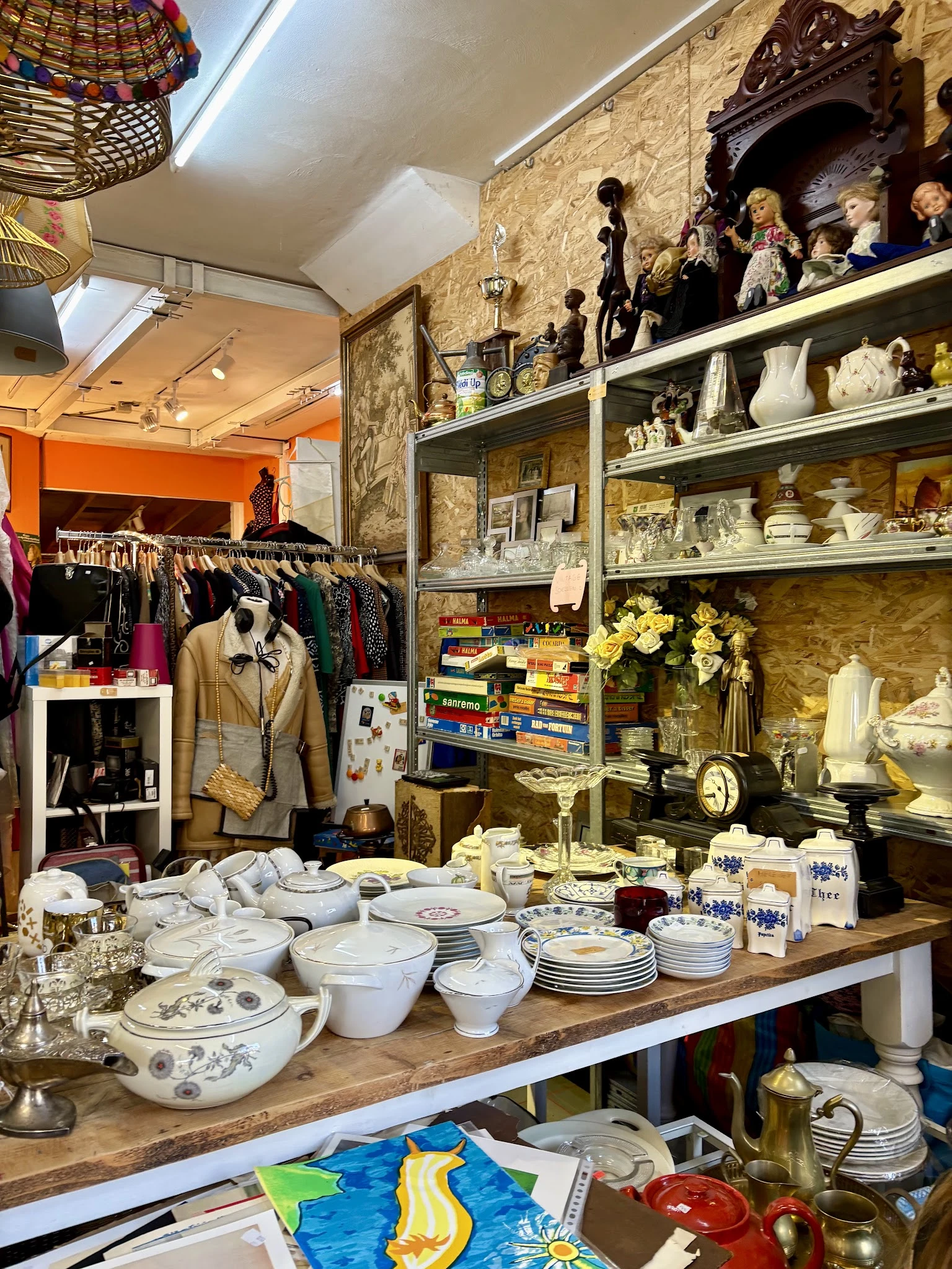 Winkelbinnenruimte met kledingrek links, open stellingen met servies en decoratieartikelen, oranje muur op achtergrond
