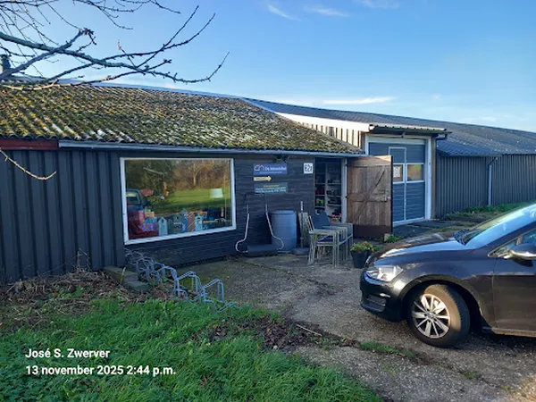Voorkant van kringloopwinkel met donkerblauwe golfplaten wanden, groot raam en parkeerplaats met auto