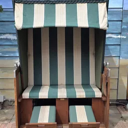 Groene en witte gestreepte strandstoel met houten armleuningen, uitgeklapt tegen een muur met strandtaferelen