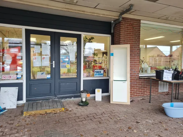 Voorgevel van kringloopwinkel met donkerblauwe deuren, etalages en ingang, bakstenen zijmuur, enkele items buiten op het terr