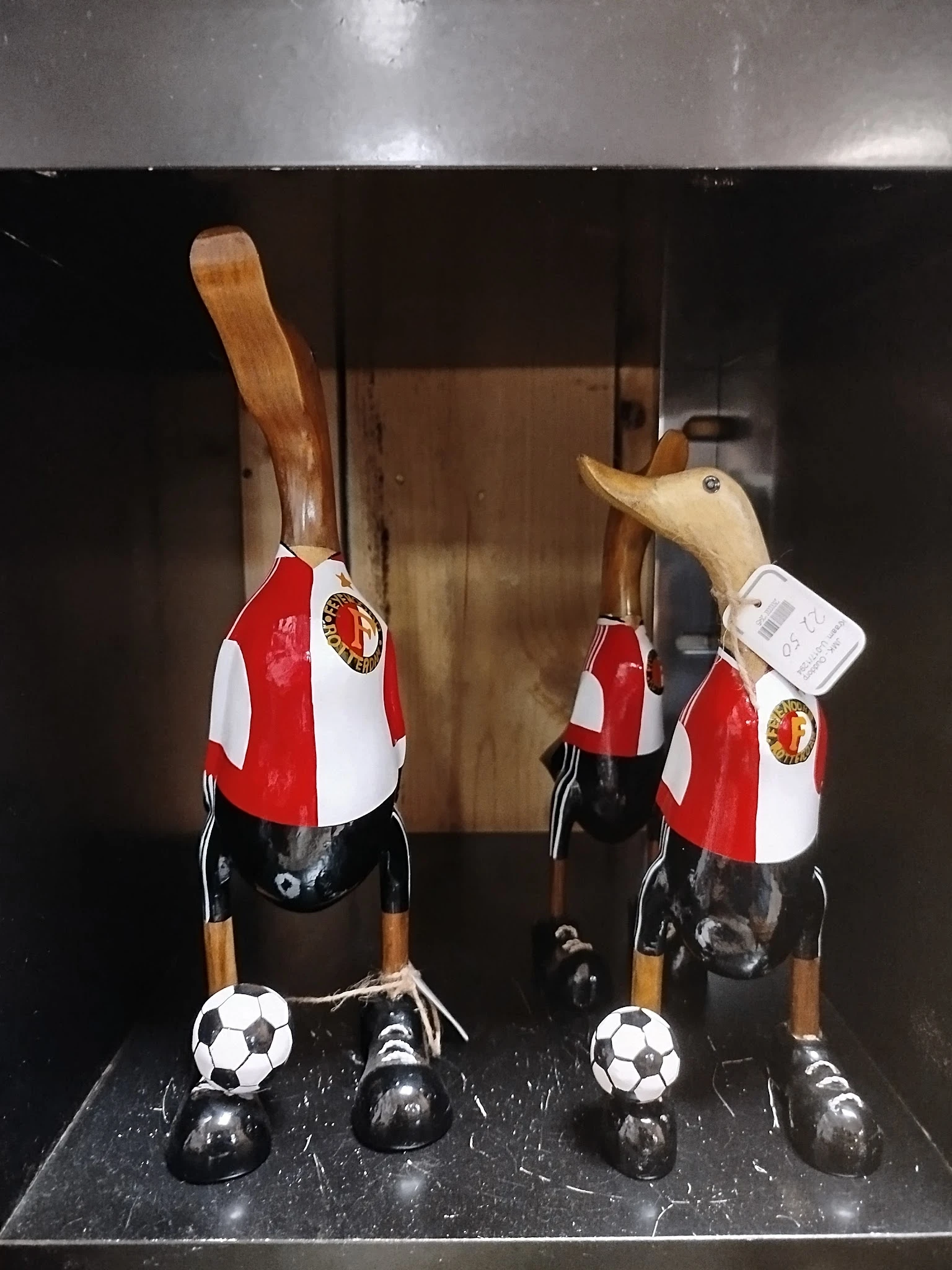 Twee rode en witte vogelhuisjes met houten dakjes en gele embleem, aan houten stokjes opgehangen in een vitrine.