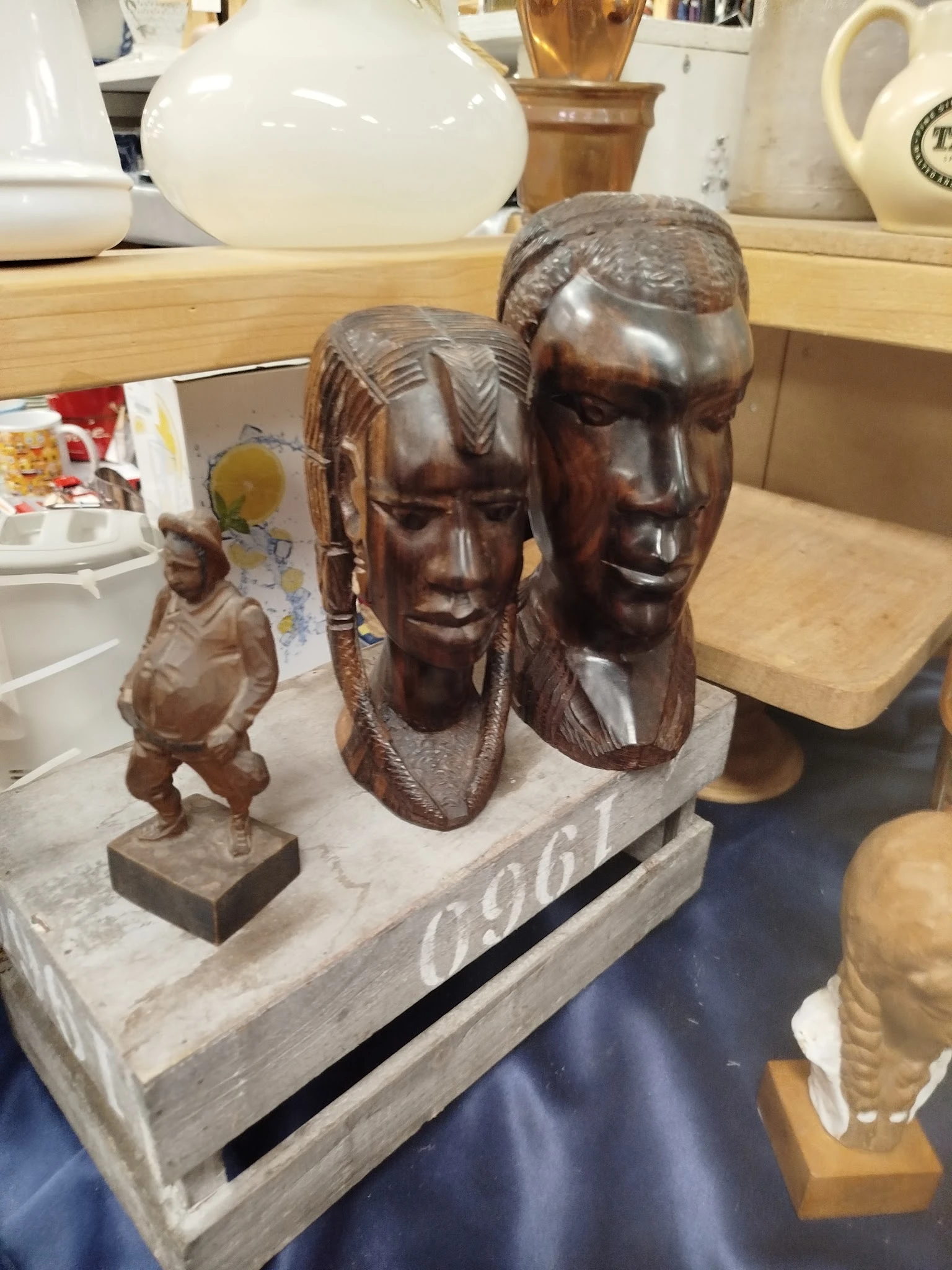 Drie houten beelden: twee grote hoofden en een kleiner figuur op een witte sokkel