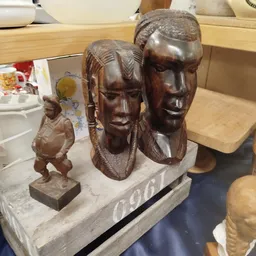 Drie houten beelden: twee grote hoofden en een kleiner figuur op een witte sokkel
