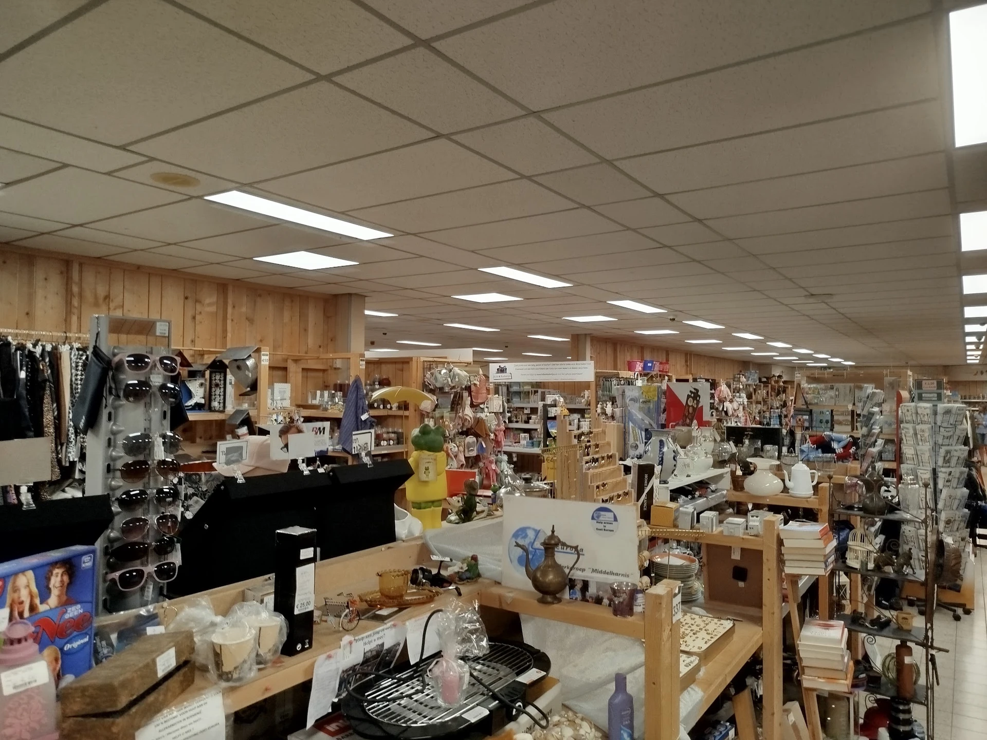 Ruim winkelbinnenland met stellingen vol tweedehands spullen, kleding en huishoudartikelen onder fluorescent licht