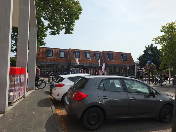 Voorkant van kringloopwinkel met rode bakstenen gevel, geparkeerde auto's op straat en fietsers op het plein