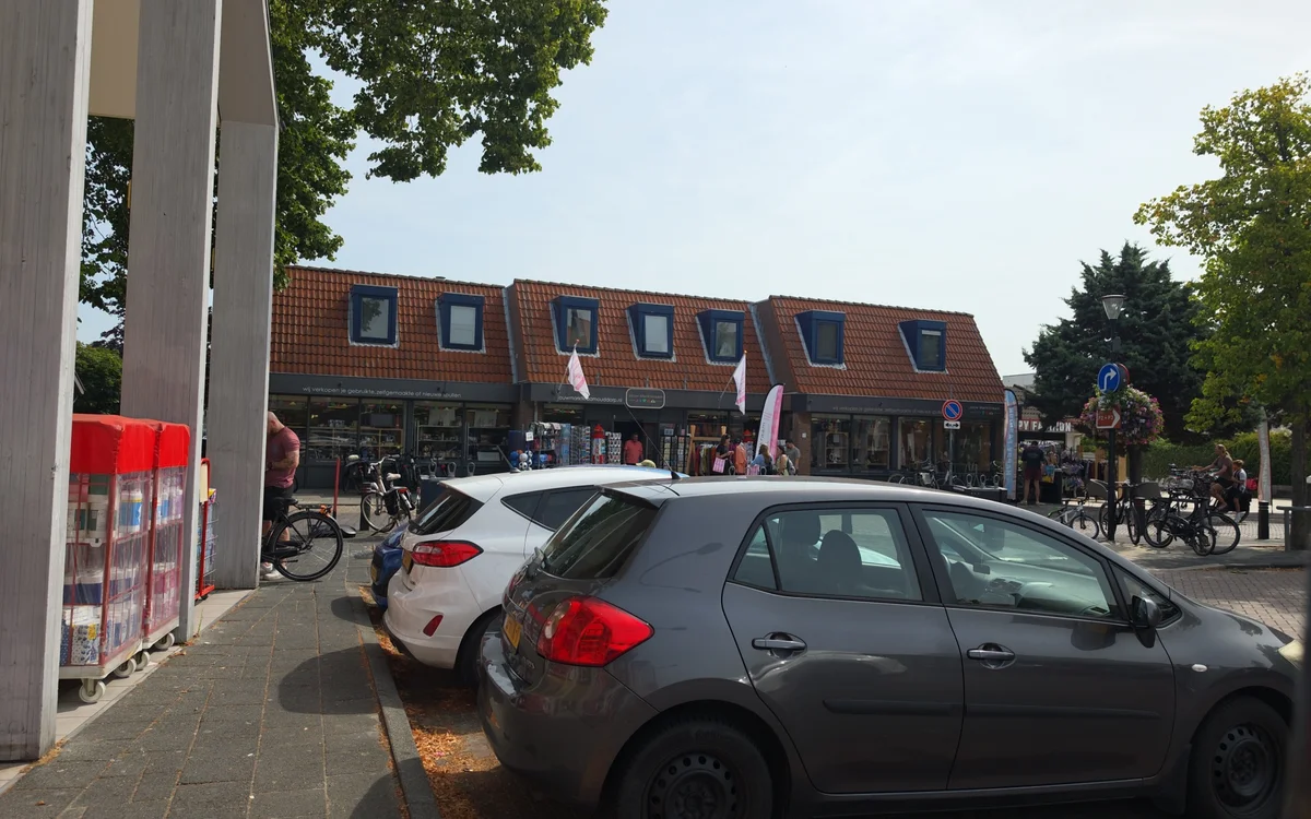 Voorkant van kringloopwinkel met rode bakstenen gevel, geparkeerde auto's op straat en fietsers op het plein