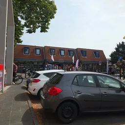 Voorkant van kringloopwinkel met rode bakstenen gevel, geparkeerde auto's op straat en fietsers op het plein