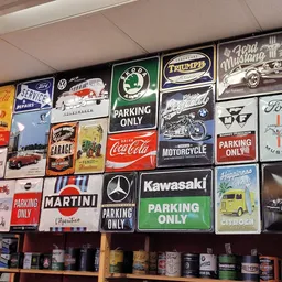Verzameling vintage emaille- en metalen reclameborden aan de muur, waaronder Coca-Cola, Ford, BMW, Triumph en Kawasaki.