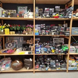 Houten stellingen volgestouwd met tweedehands spullen: bloemen, dozen, elektronische apparaten, keukenspullen en ander huisho