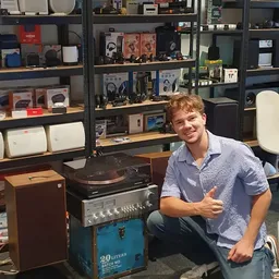 Jonge man posant naast vintage audio-apparatuur in een kringloopwinkel met stellingen vol elektronicaproducten op de achtergr