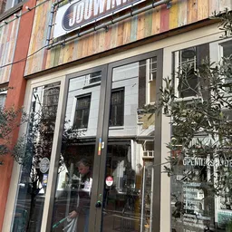 Glazen pui van kringloopwinkel met groene frame en planten aan beide zijden van de ingang