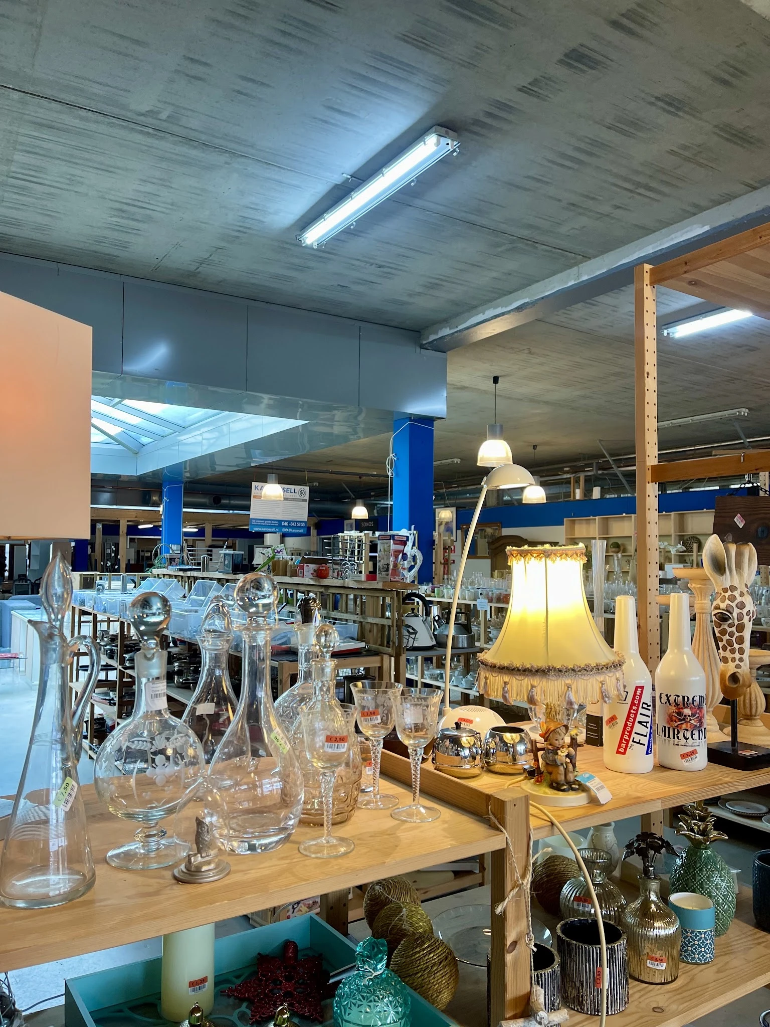 Uitgestalde huishoudelijke artikelen en decoratie in de kringloopwinkel, waaronder glazen, lampen en keramiek op houten tafel